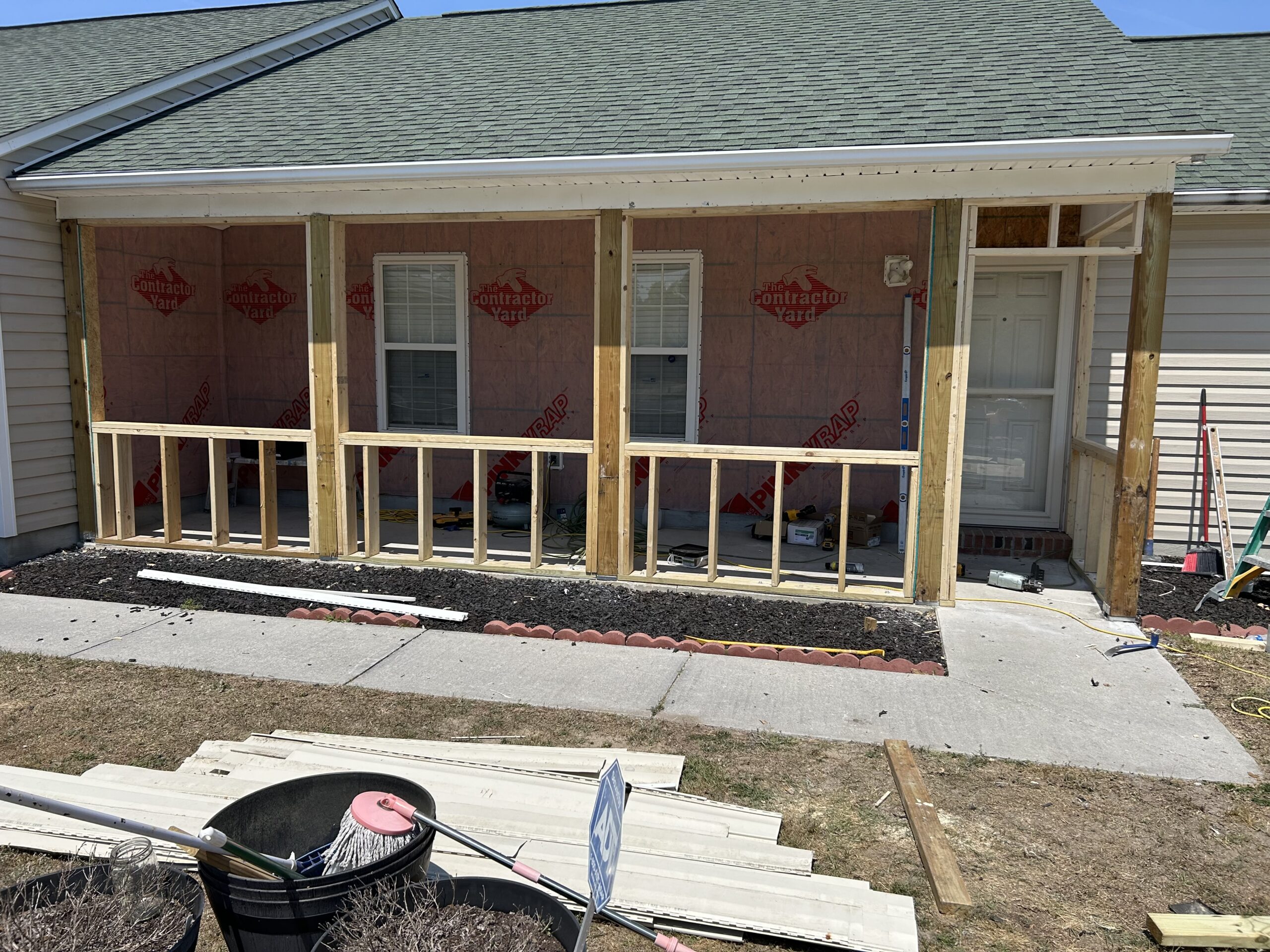 A house porch is under construction with wooden framing and insulation visible, tools and materials scattered, and the yard partially landscaped.