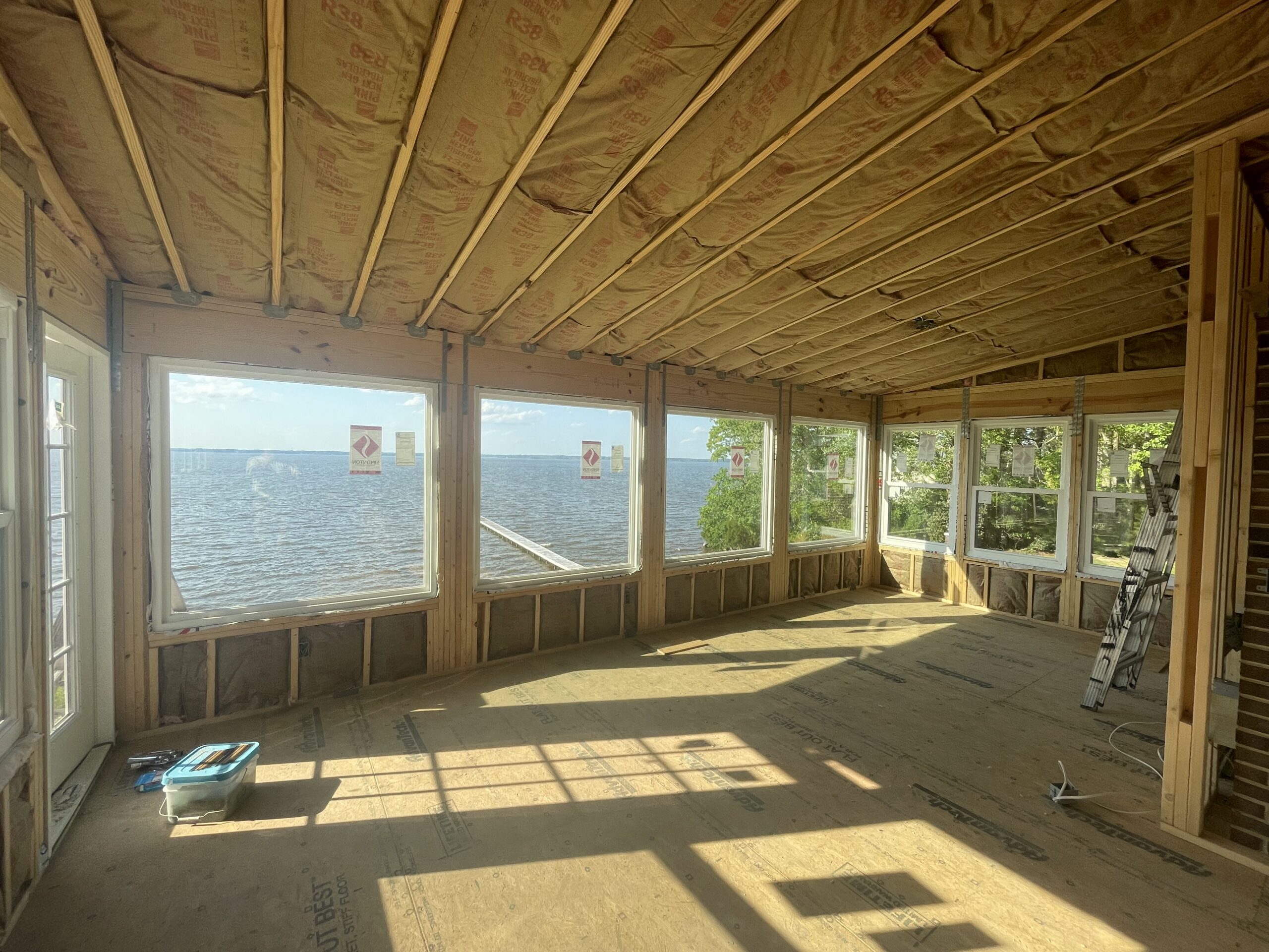 Room under construction with exposed insulation and wooden framing, facing large windows with a view of a body of water and trees outside. Construction materials are visible on the floor.