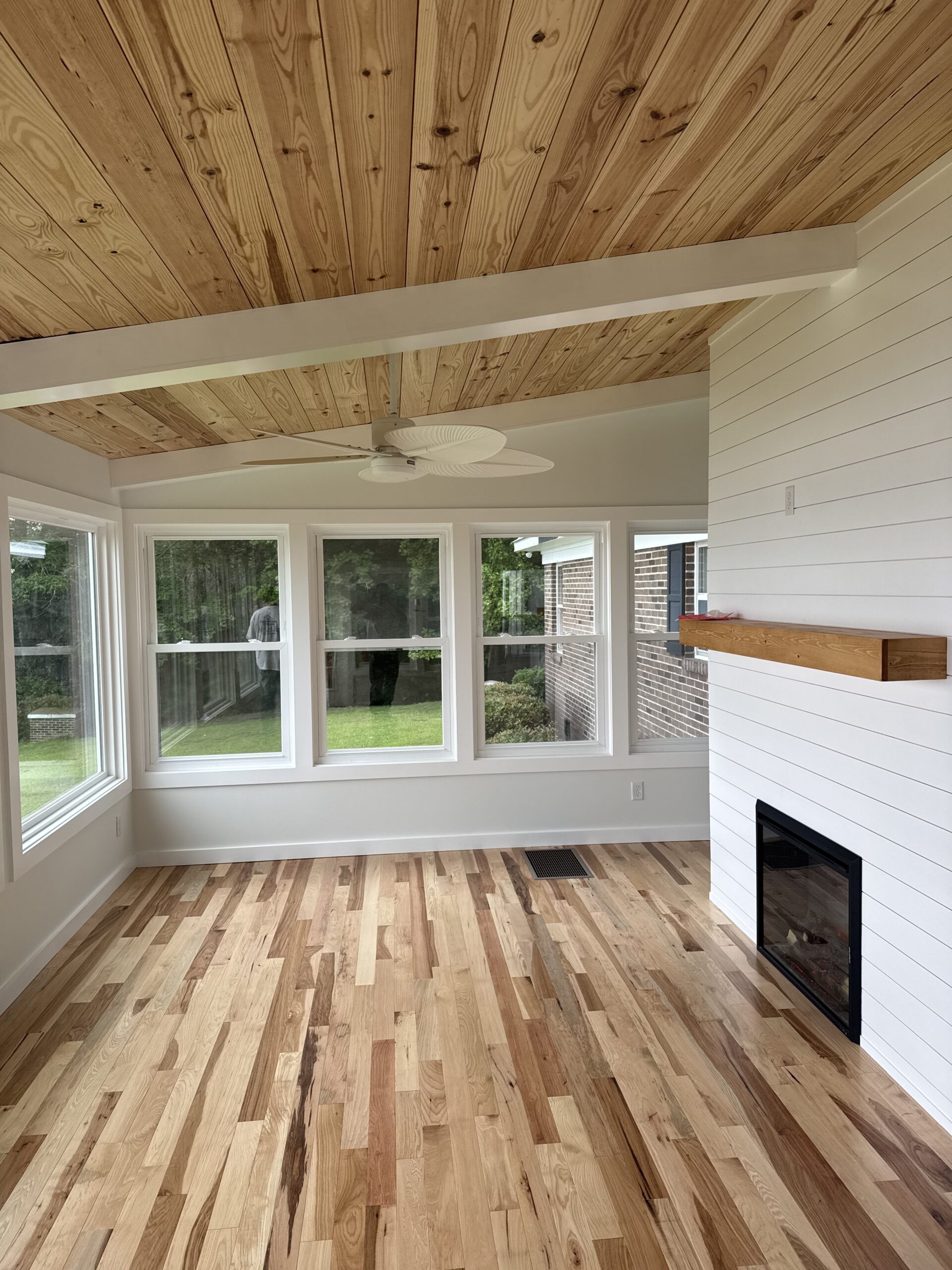 Bright sunroom with large windows, light wood floor, shiplap walls, wood ceiling with exposed beams, ceiling fan, and a wall-mounted fireplace with a wooden mantel.