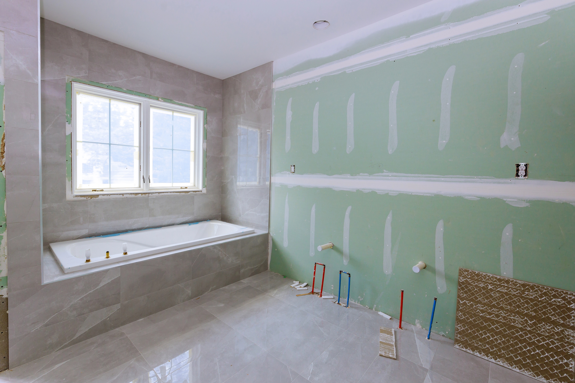 Bathroom under construction with a bathtub installed, unfinished drywall, exposed plumbing, and tile pieces on the floor.
