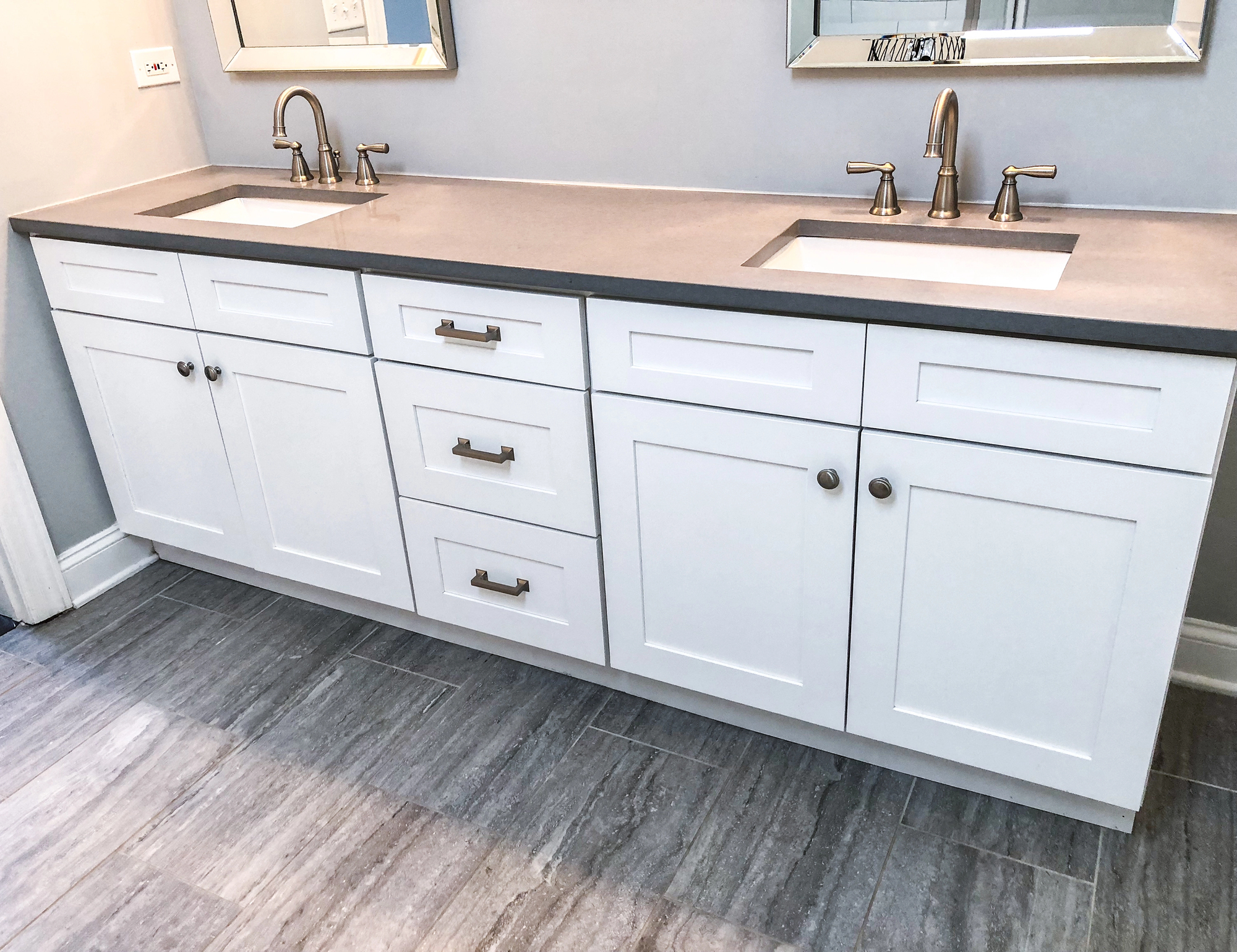 Double-sink bathroom vanity with white cabinets, gray countertop, and brushed metal fixtures, set against a light gray wall with tile flooring.