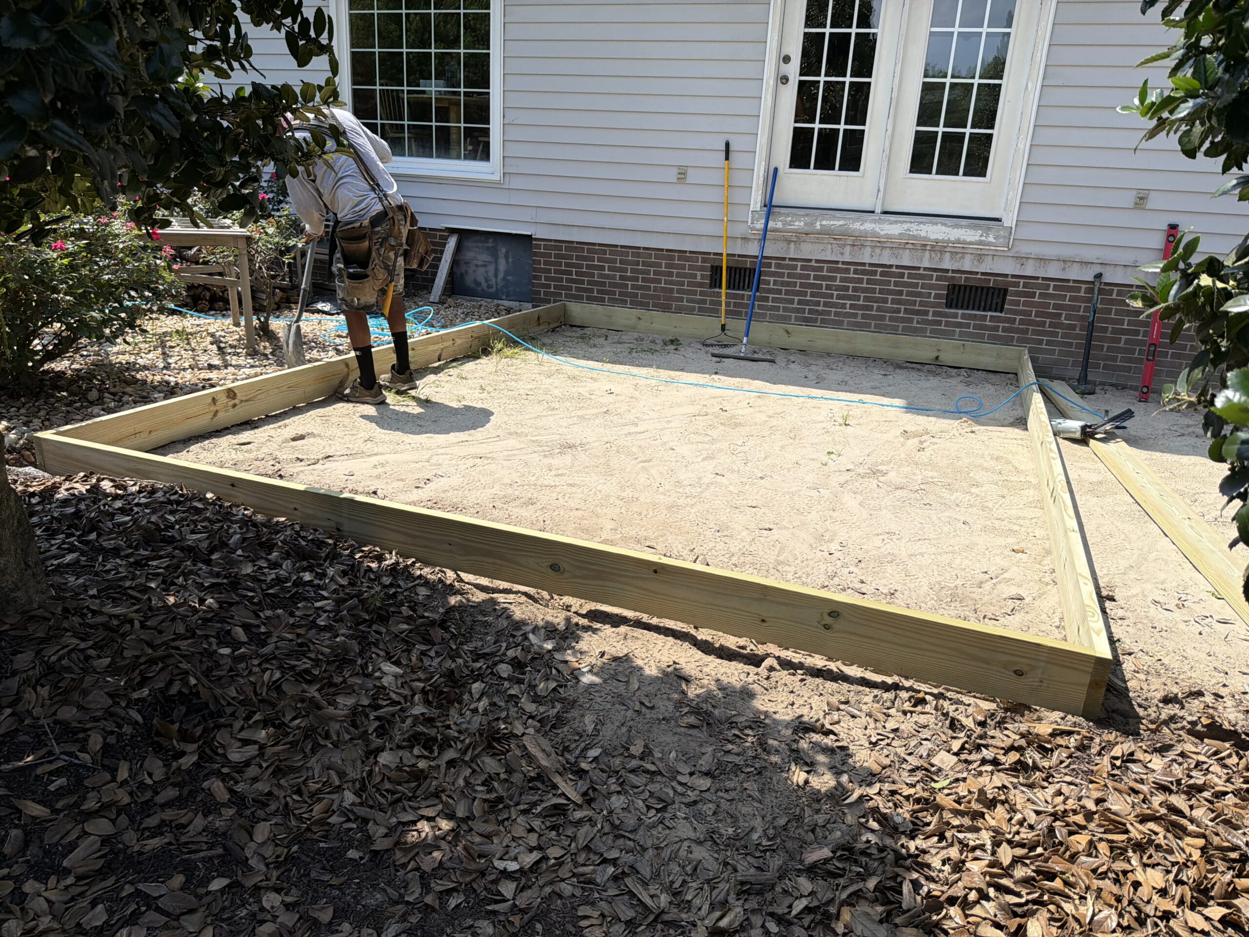 A person stands on sand inside a wooden frame, preparing a foundation next to a house with white siding and double glass doors. Tools and leaves are visible nearby.