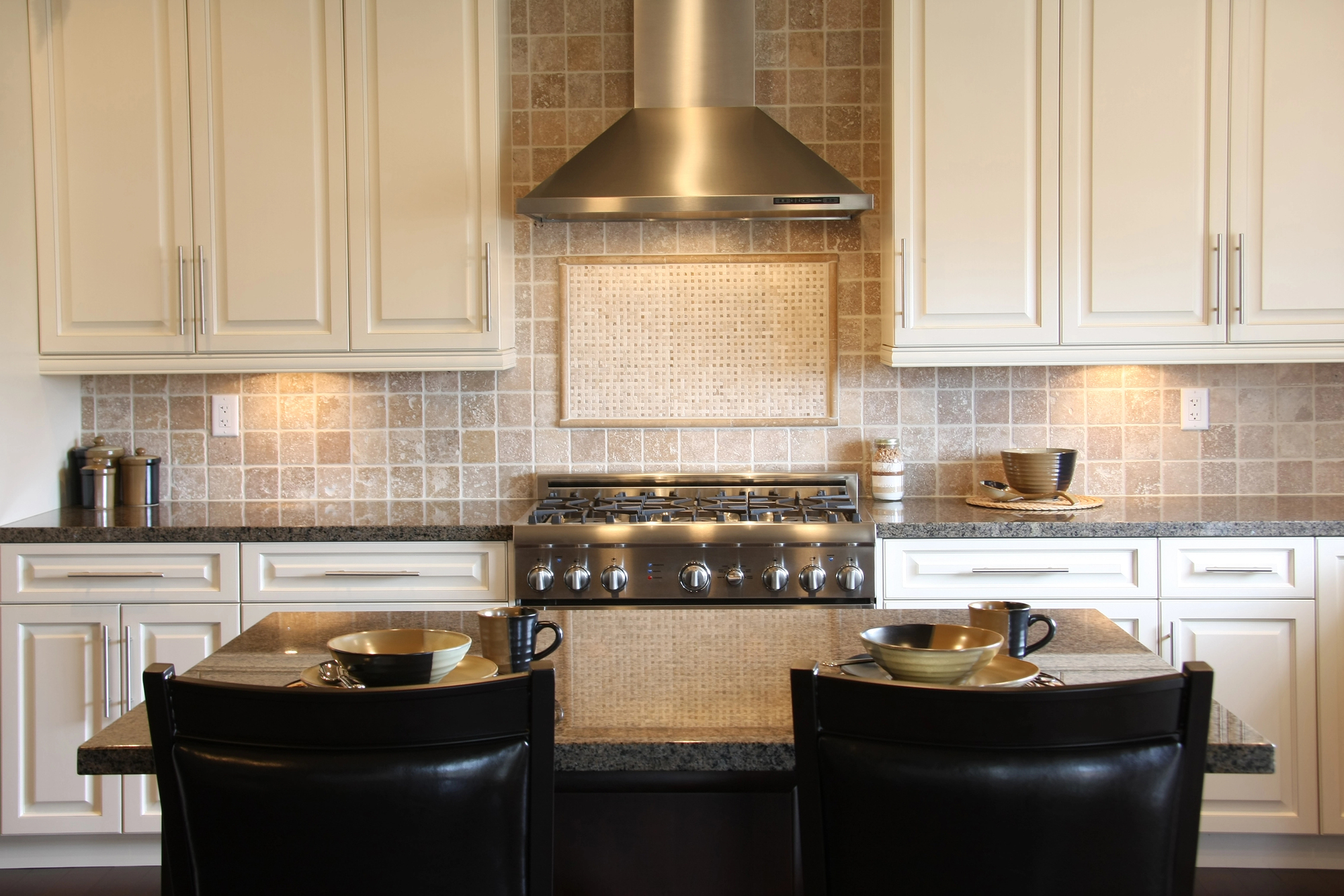 Modern kitchen with stainless steel stove and hood, beige tile backsplash, white cabinets, and a granite island set with two bowls and mugs; two black chairs face the island.
