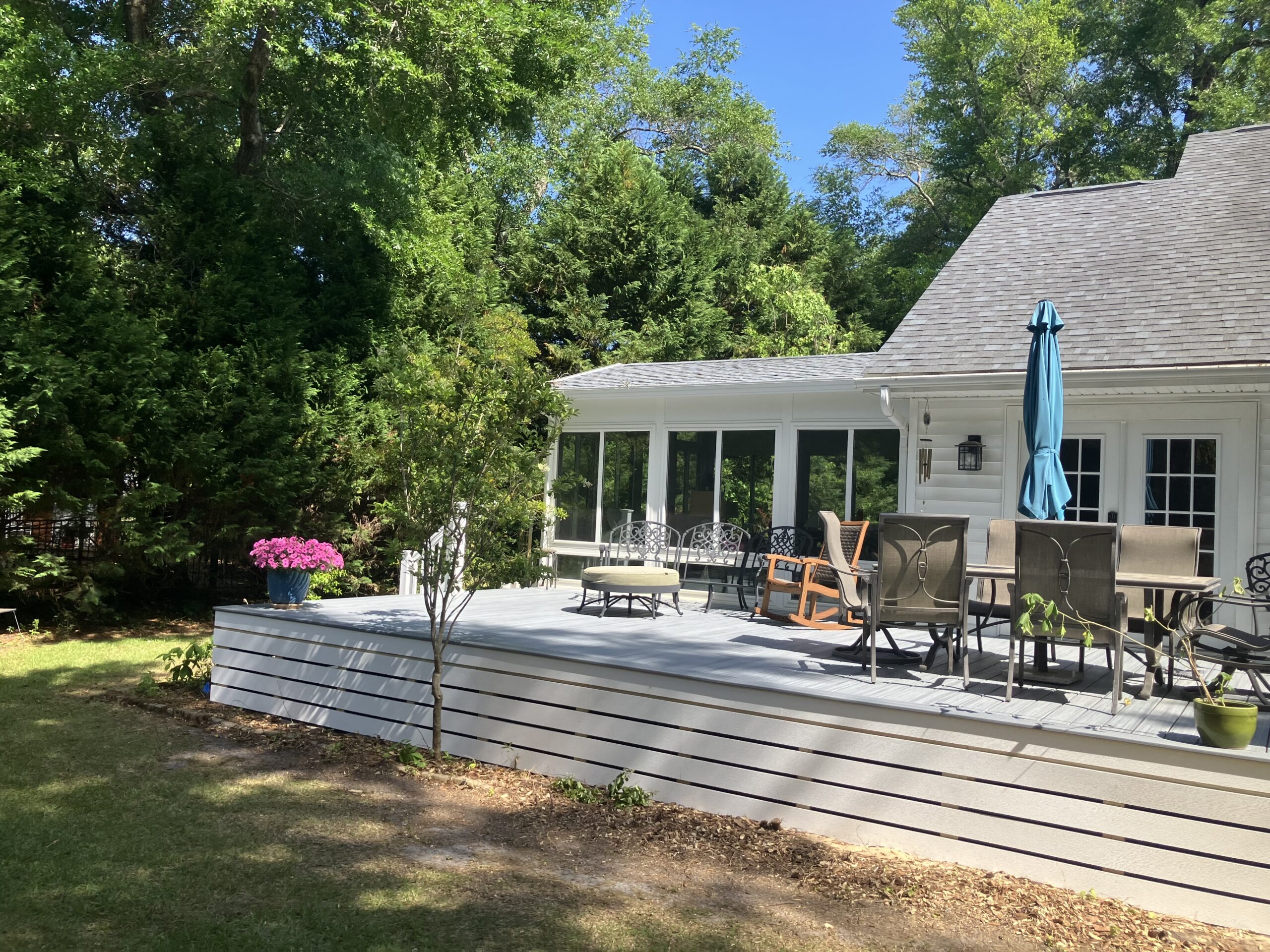 A white house with a gray deck, outdoor furniture, potted plants, and a blue umbrella, surrounded by trees and greenery on a sunny day.