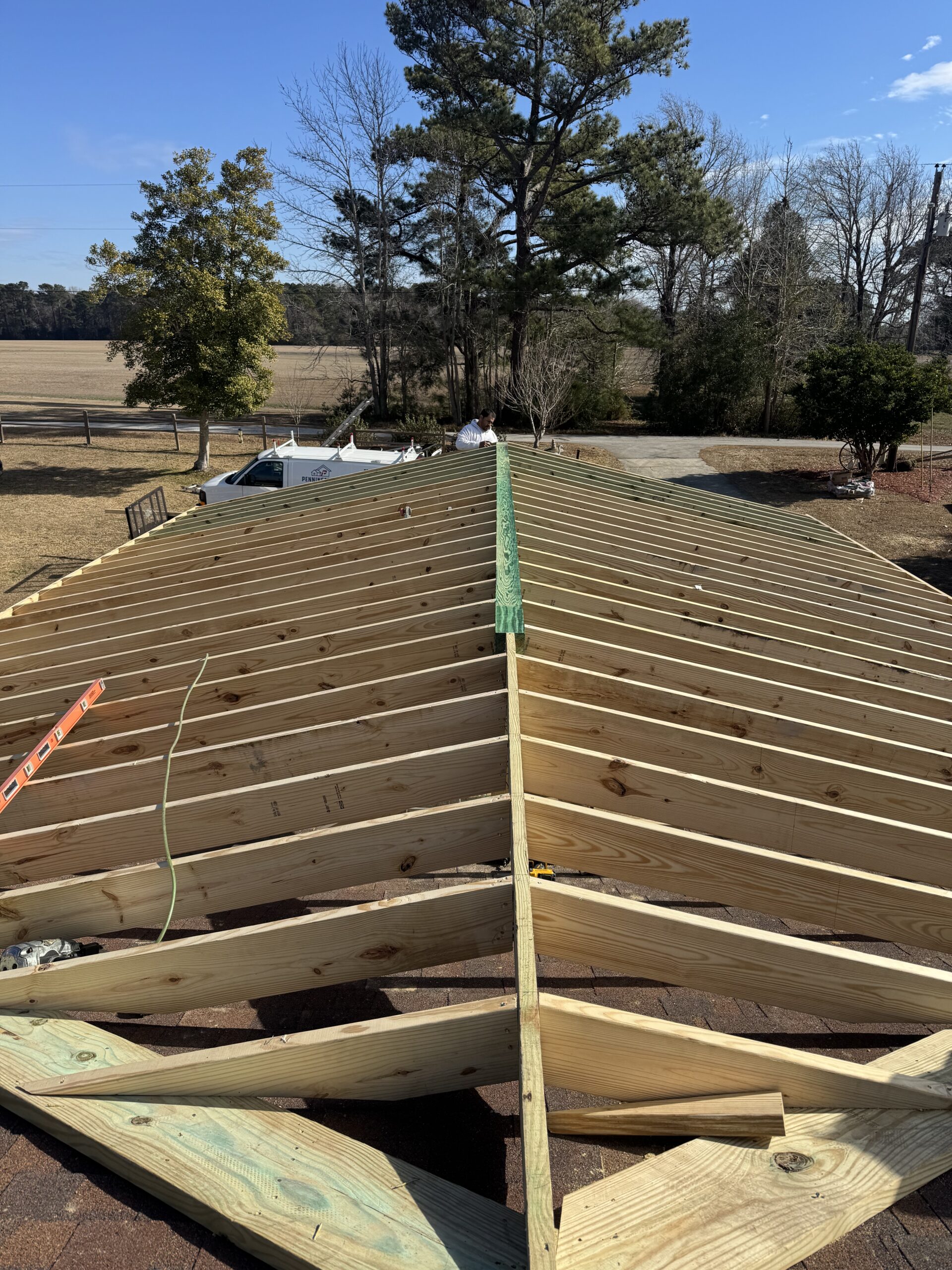 View of a partially constructed wooden roof frame with exposed beams, ladders on the side, and trees and fields in the background under a clear sky.