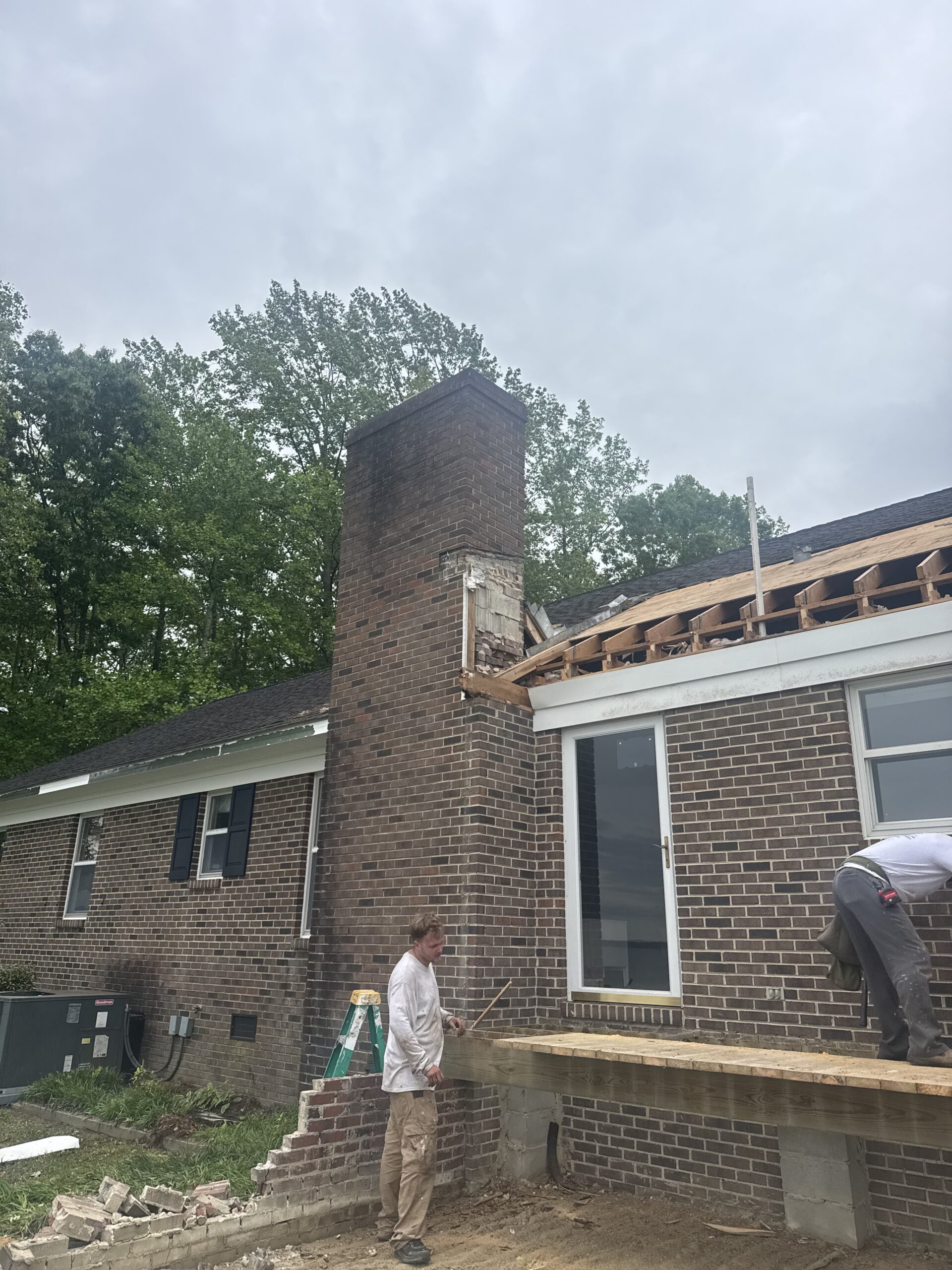 Two workers are renovating a brick house, with part of the roof and chimney under construction. Debris and construction materials are visible on the ground.