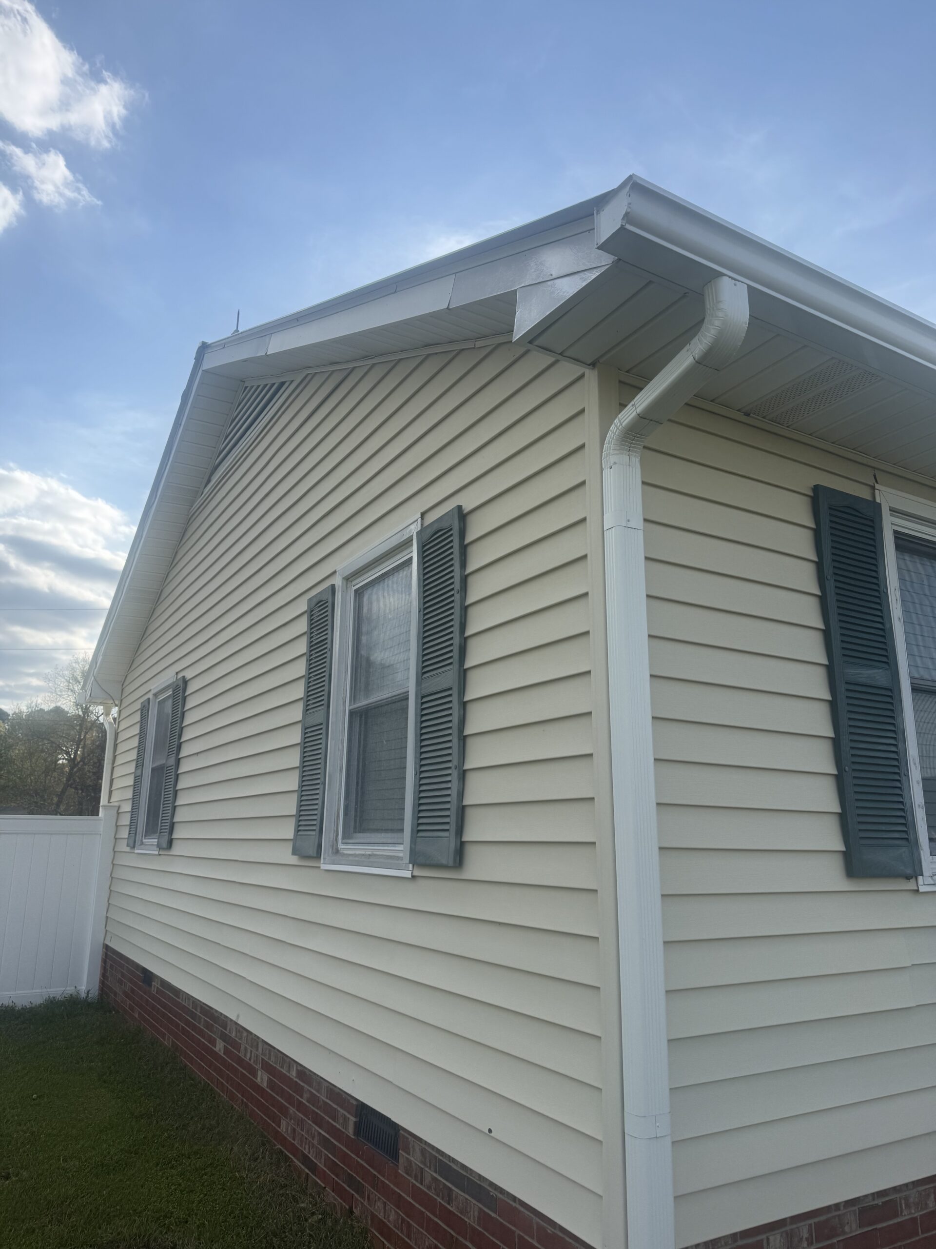 A beige house with green shutters, white gutters, vinyl siding, and a red brick foundation is shown under a partly cloudy sky.
