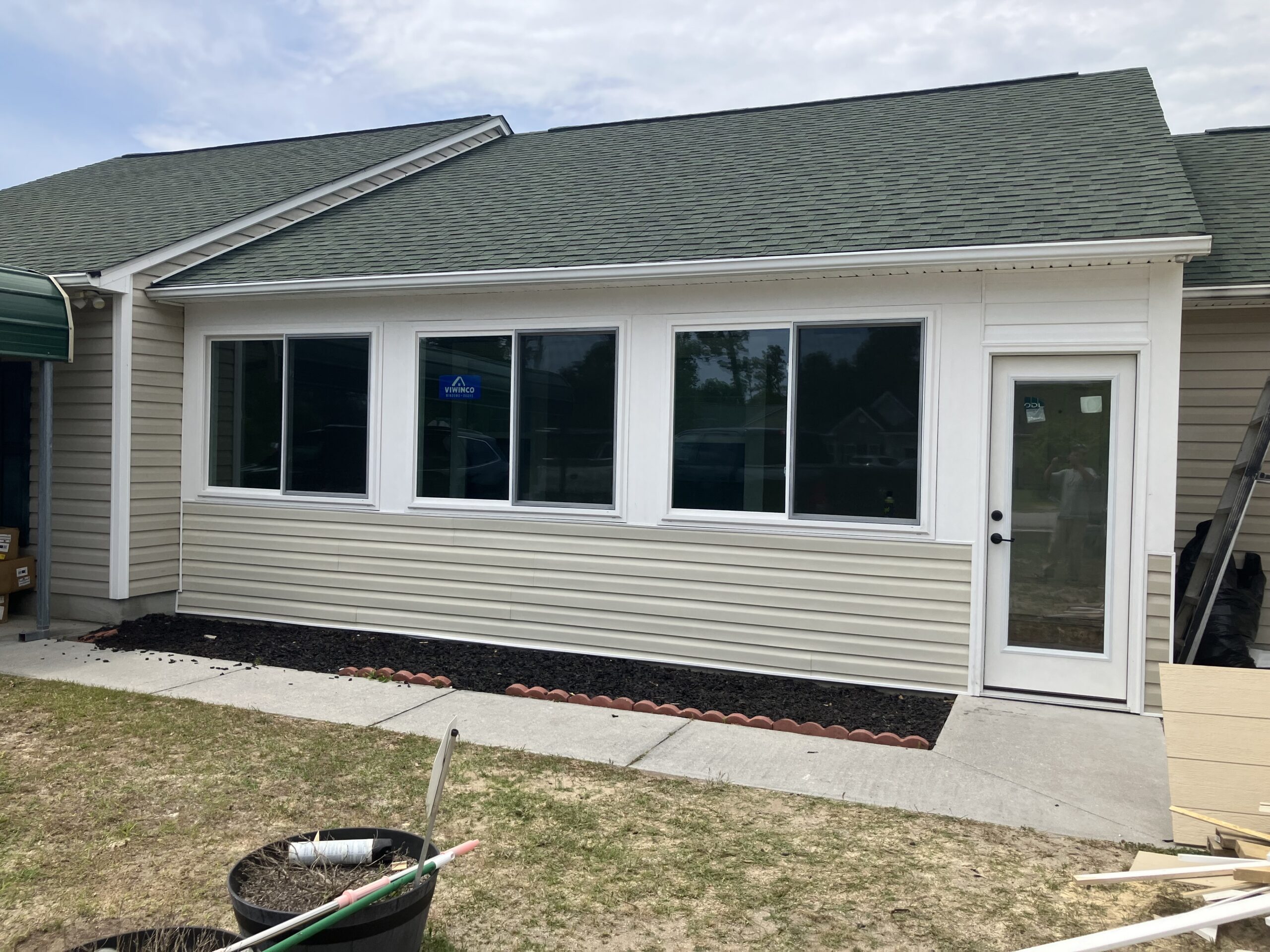 A newly constructed sunroom with large windows and a white-framed door, attached to a house with green shingles and beige siding. Construction materials are visible in the foreground.