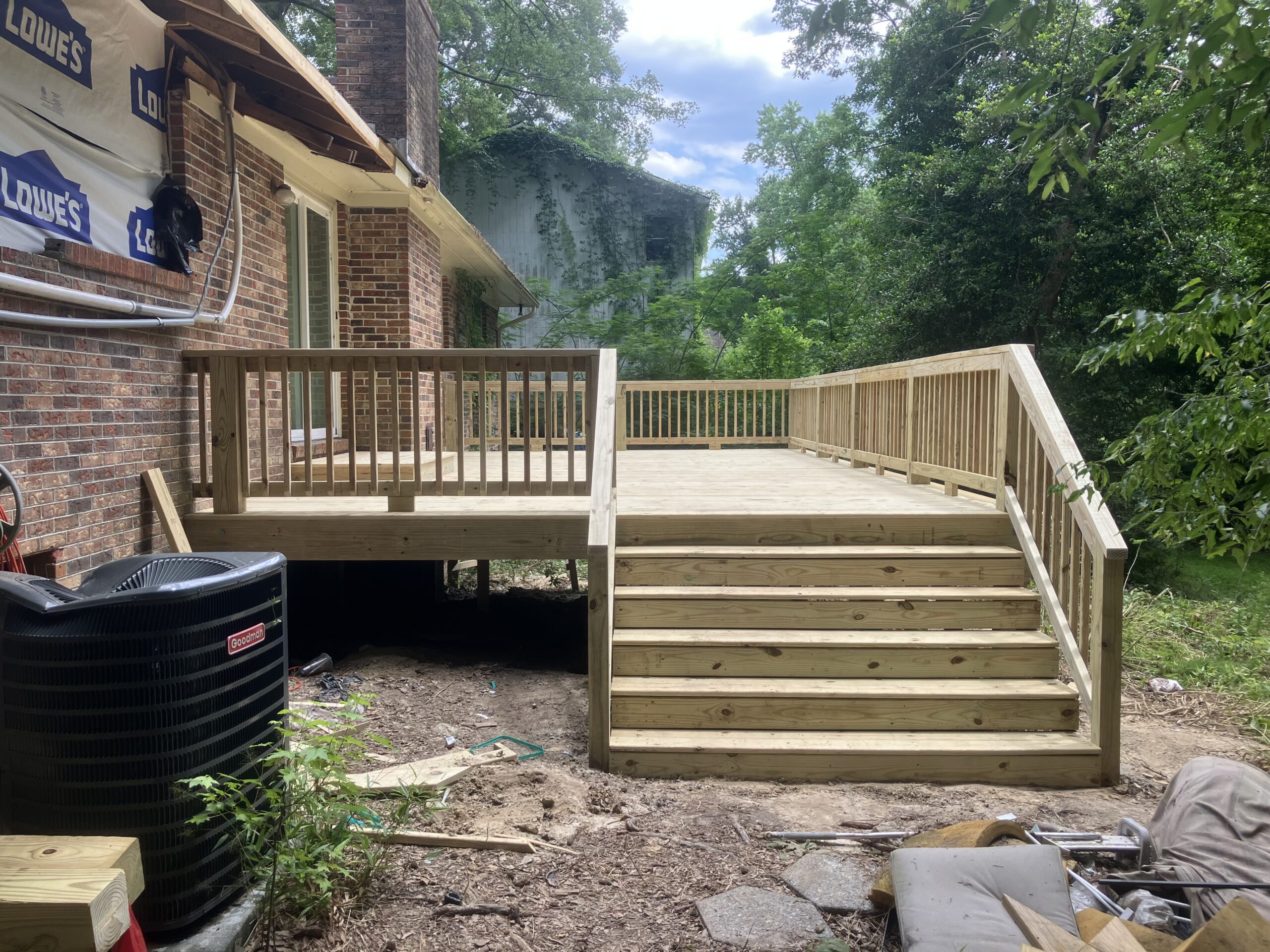 A newly built wooden deck with stairs and railings attached to a brick house, overlooking a backyard with trees and an HVAC unit nearby.