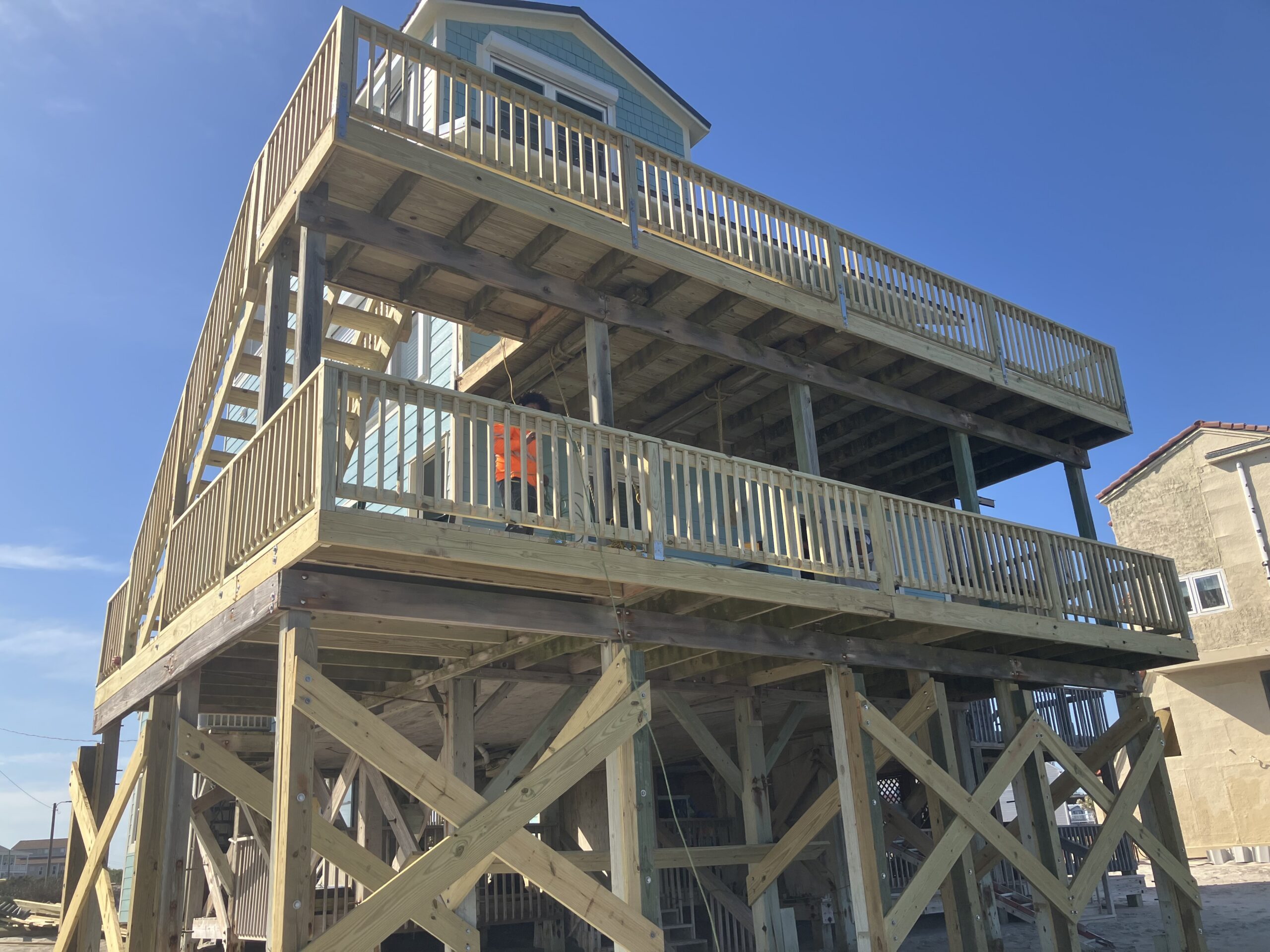Two-story wooden beach house elevated on stilts, with large wraparound decks on both levels and a clear blue sky in the background.