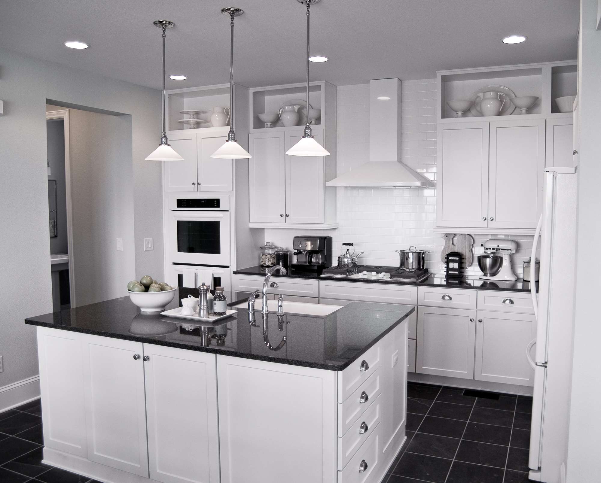 Modern white kitchen with an island, black countertops, stainless steel appliances, hanging lights, and open shelves displaying dishes and bowls.
