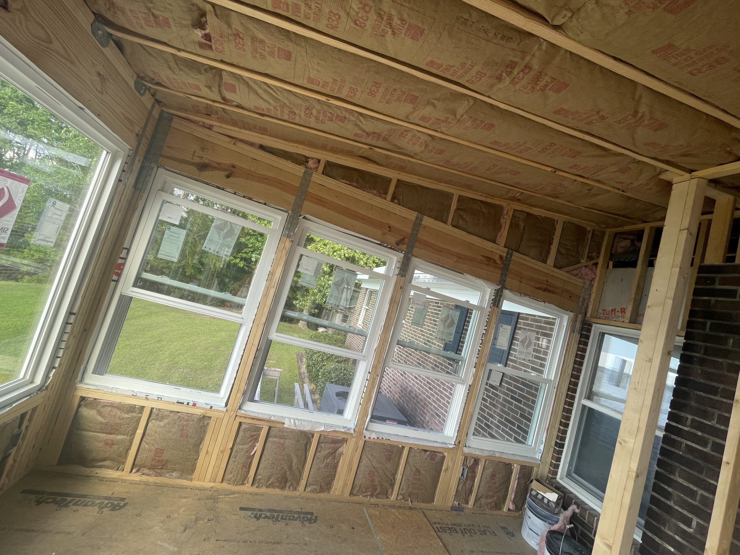 A partially finished sunroom with exposed insulation, wooden framing, and a row of large, newly installed windows facing a green lawn.