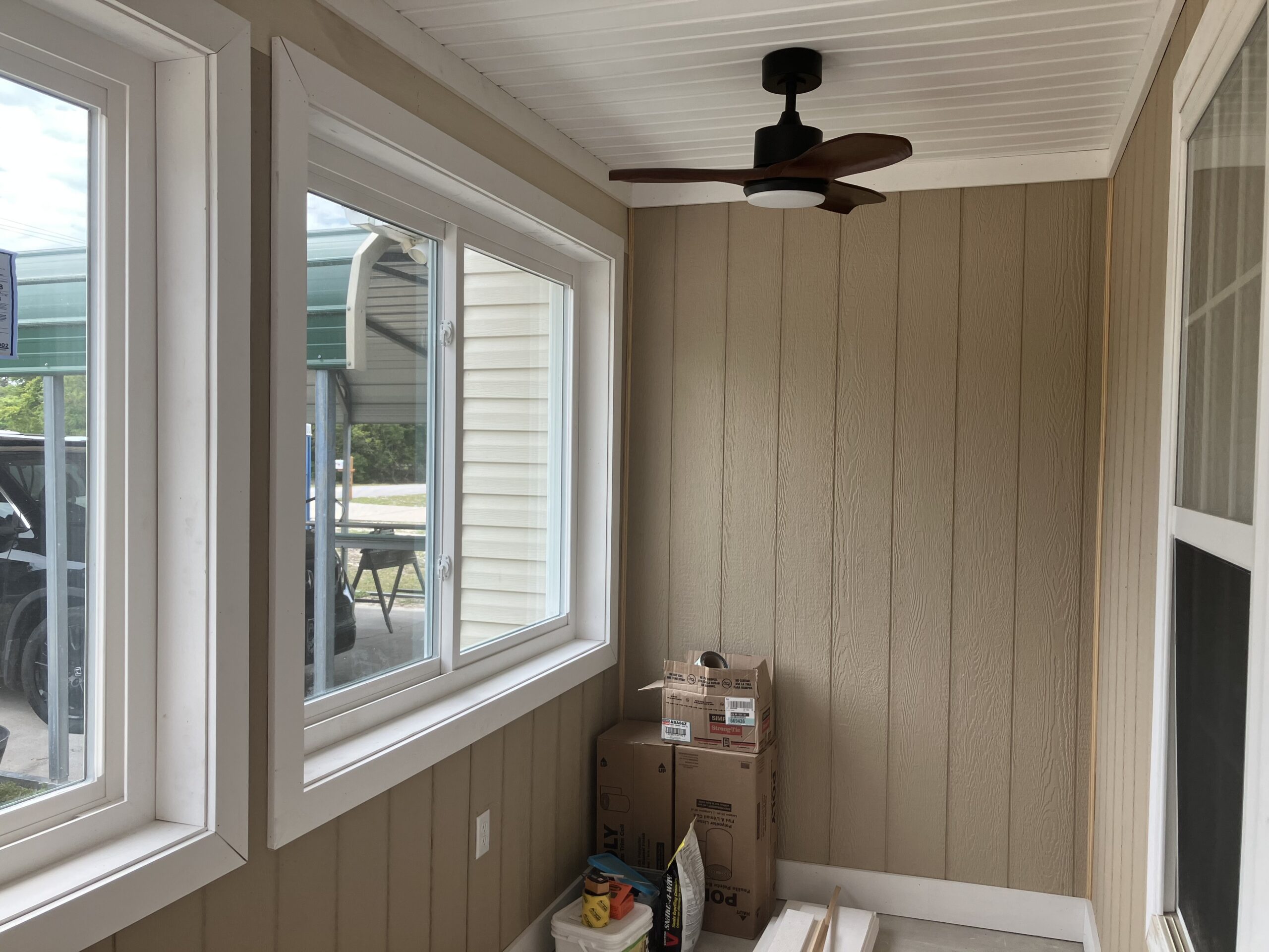 A small enclosed porch with tan vertical paneling, a ceiling fan, two windows, and several boxes and items stacked in the corner.