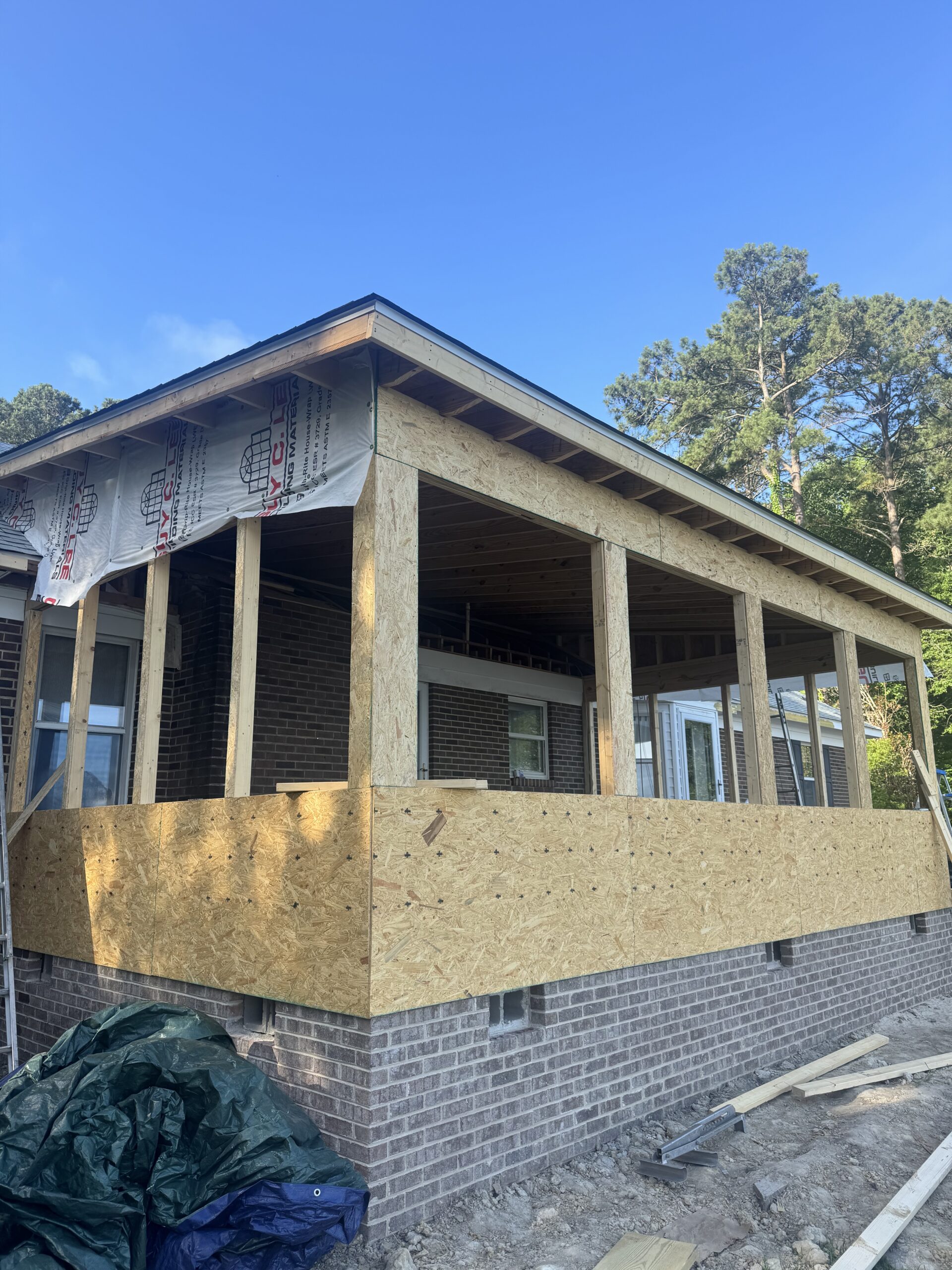 A house under construction with plywood and framing added to create a new covered porch area. Construction materials are visible on the ground.